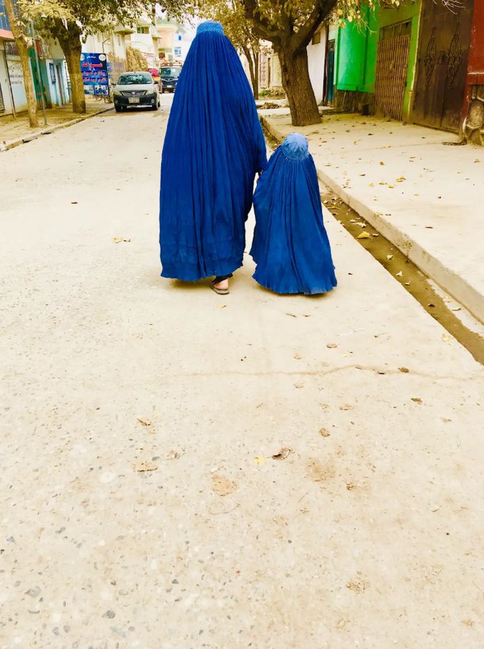 Two people of different heights wearing blue burqas walk together along a tree-lined street.