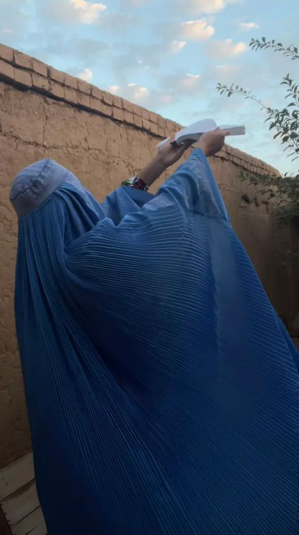 A person in a blue burqa holds an open book toward the sky beside an earthen wall.